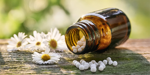 Braune Glasflasche liegt auf Holzoberfläche, weiße Tabletten fallen heraus. Um die Flasche liegen mehrere weiße Gänseblümchen mit gelber Mitte. Hintergrund unscharf mit grünen Farbtönen.