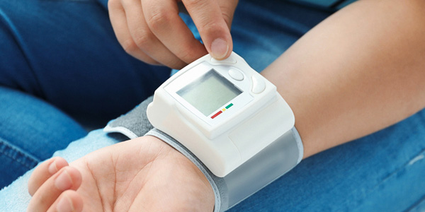 Nahaufnahme eines Handgelenks mit digitalem Blutdruckmessgerät. Die Person drückt eine Taste am Gerät. Das Display zeigt Messwerte und Symbole. Das Gerät ist mit einem Band am Handgelenk befestigt.